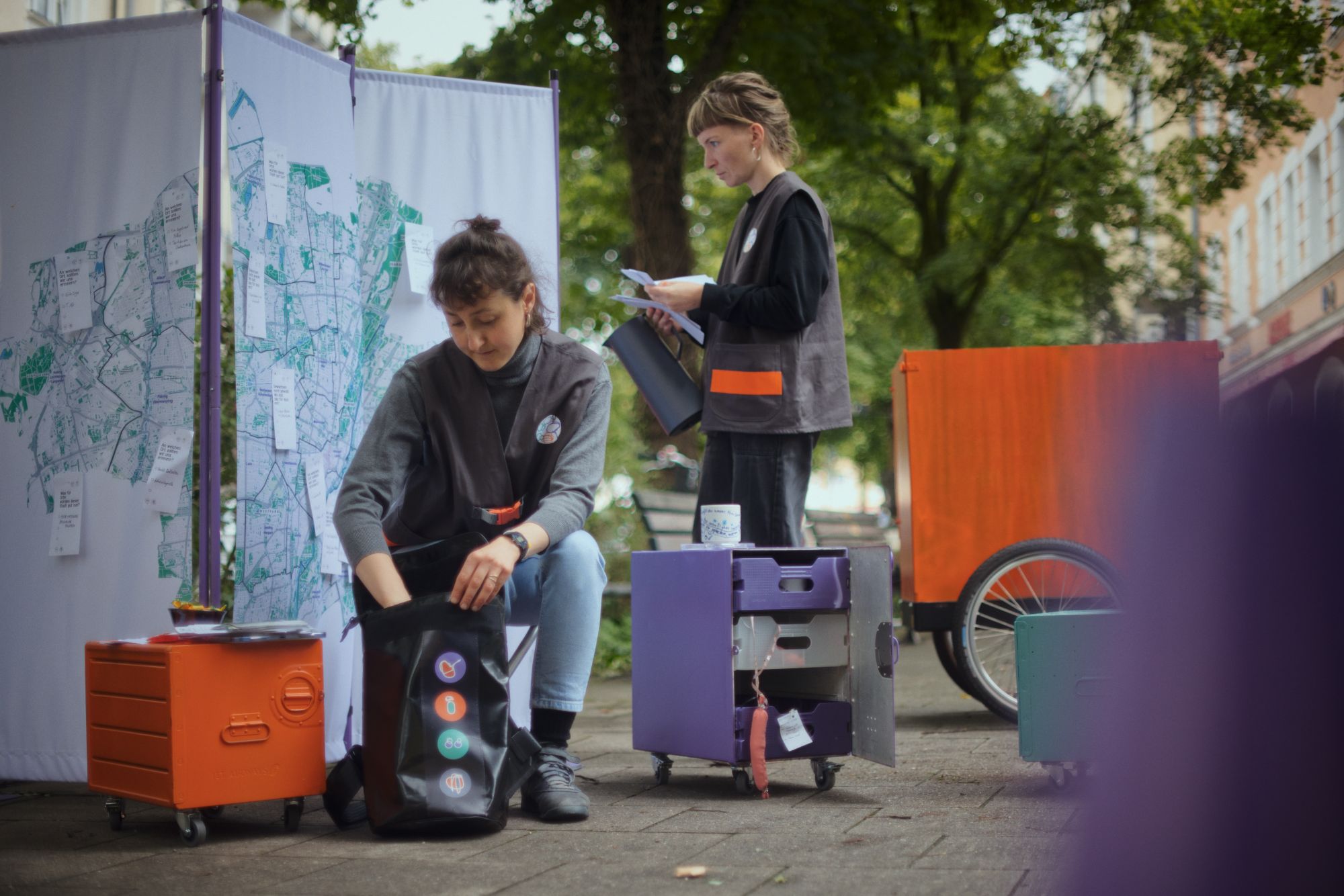 Ein Paravan mit Stadtkarte im öffentlichen Raum, davor zwei junge Frauen in Westen. Der Forschungsrucksack wird gepackt und es stehen rollbare Flightcases herum (orange und lila).